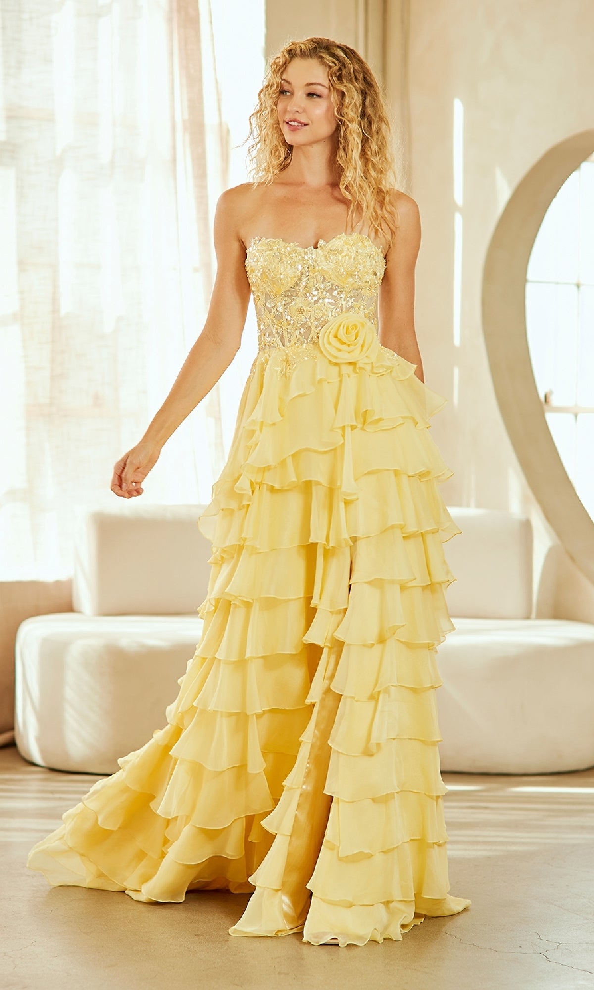 A woman stands indoors in soft light wearing the Strapless Ruffled Prom Gown: Amelia Couture 7087, featuring a sequined bodice, ruffled skirt, and floral detail.